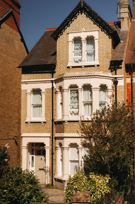 A multi-storey Victorian-style house made of yellow brick with white decorative window frames and sills, situated on a residential street. The building features a gabled roof with dark shingles, and b
