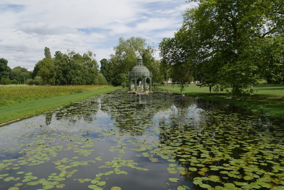A classical stone garden pavilion with tall, fluted columns supporting a triangular pediment, situated amidst lush greenery and vibrant flowering plants, with stone steps leading up to its entrance. T