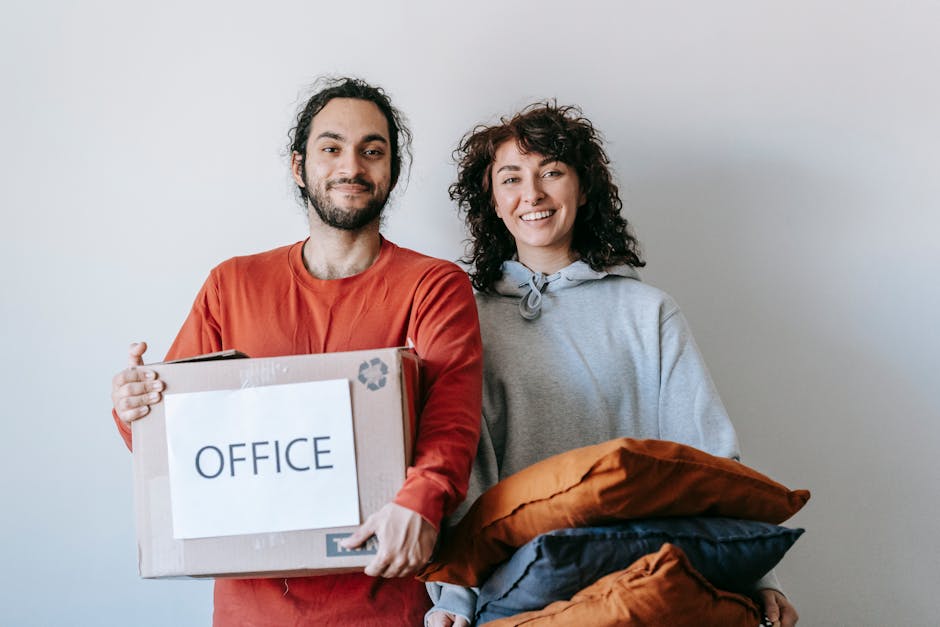 A man and a woman standing side by side against a plain light-colored wall during a home relocation, with the man holding a cardboard box labeled 'OFFICE' in bold letters. The box is wrapped with packing tape and appears to contain office items or documents. The woman is smiling and holding a bundle of soft fabric items, such as cushions or clothing, in her arms. Behind them, there are no visible windows or furniture, but the scene suggests they are preparing for a furniture transport or moving process. The bright, well-lit environment indicates ambient lighting, and both individuals appear relaxed, representing the packing and loading stage of a house or office move, related to professional removals services by Chiswick Movers.