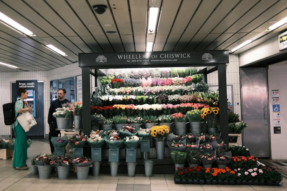 A flower stall named 'Wheeler of Chiswick' situated in an underground station with a variety of colorful flowers organized in metal buckets and containers on multiple levels. The display includes roses, sunflowers, and various other potted plants, arranged neatly against a black backdrop with a sign above. The station has a tiled ceiling with fluorescent lighting and tiled walls. To the left, a woman with a backpack and light-colored trousers is engaging in conversation with a staff member dressed in dark clothing. Behind them, an ATM and a sign indicating cash withdrawals are visible. The scene captures a moment of home relocation or moving logistics involving loading or unloading flowers in a busy urban environment, with the flowers and display point-of-sale being suggestive of packing and transport elements relevant to house removals or commercial relocations managed by Chiswick Movers.