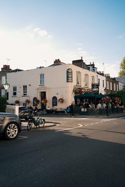 A street view of a white, two-story building with multiple windows and flower baskets hanging beneath the windows and near the entrance, which appears to be a pub or restaurant, with an outdoor seating area protected by a dark green awning. Several people are seated at tables in the outdoor area, and pedestrians are walking along the sidewalk. Parked cars and bicycles are visible in front of the building, with a clear blue sky and a few clouds above. The scene depicts a lively urban environment typical of a charming neighbourhood such as Turnham Green, where professional removals and home relocations by Chiswick Movers are often coordinated, including furniture transport and packing logistics involving vehicles and loading areas.
