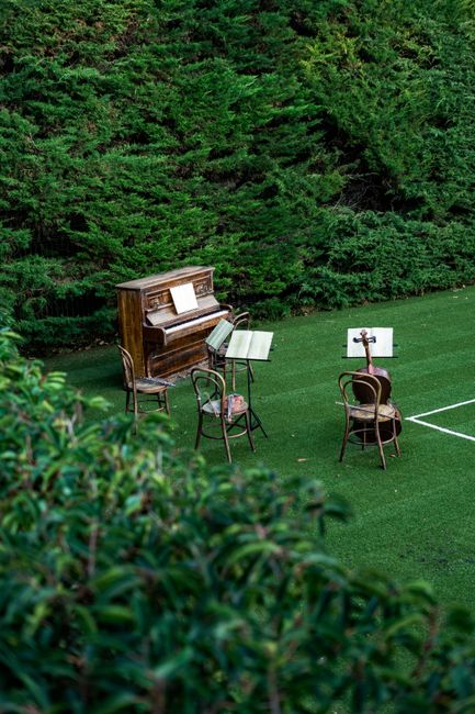 A set of three wooden chairs, a vintage upright piano, and two music stands are arranged on a well-maintained green lawn, with the piano positioned against a background of dense, tall green hedges or trees. The chairs are placed in front of the music stands, which hold open sheet music. The scene appears to be outdoors, possibly in a garden or park area, and the arrangement suggests a musical performance or practice session. The lighting is natural, indicating daytime, and the setting provides a peaceful environment suitable for music-related activities. No moving activity or additional objects are visible, and the scene is captured from a slightly elevated angle, focusing on the musical instruments and furniture on the lawn.
